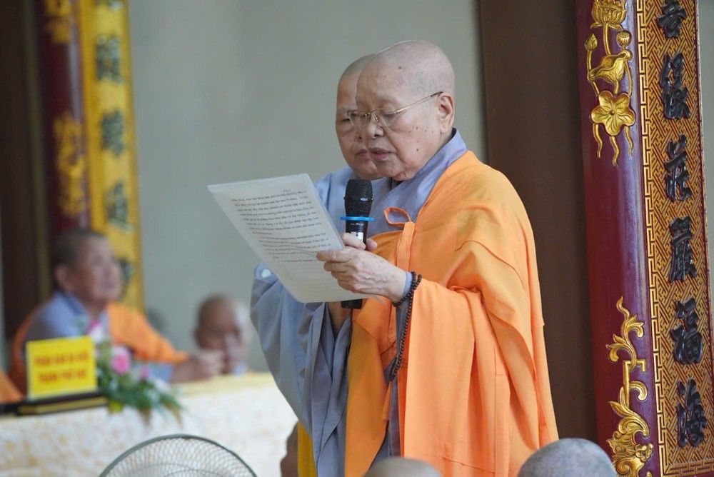 A person in an orange robe holding a microphone and reading a paper

AI-generated content may be incorrect.