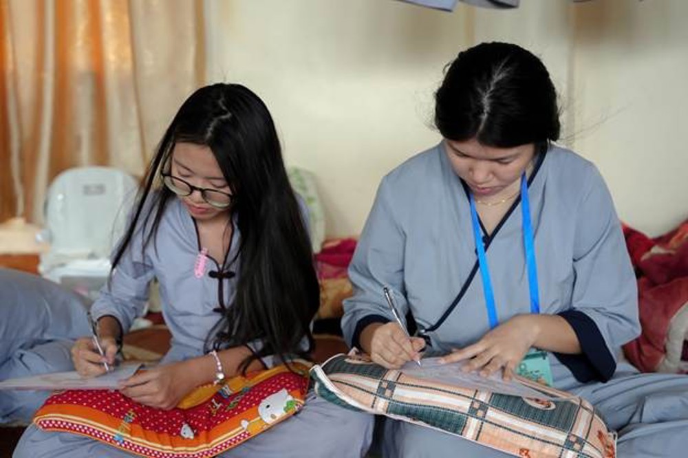 A few women sitting on a bed writing on a piece of paper

AI-generated content may be incorrect.